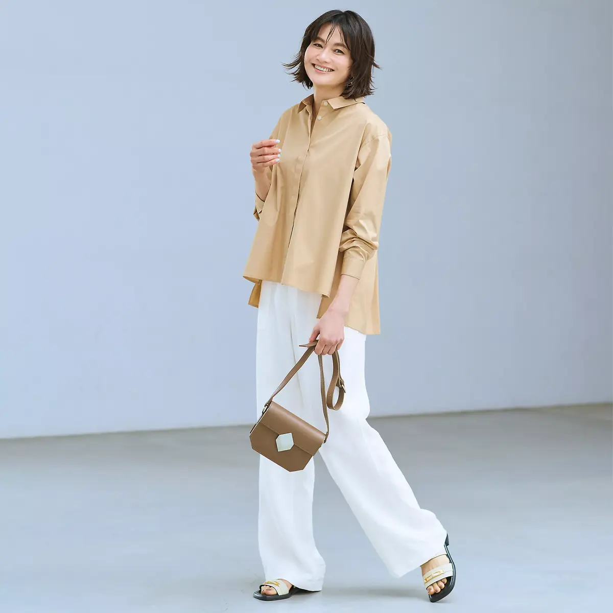 ベージュのシャツに白いワイドパンツを着用した女性が、笑顔でこちらを見ている。茶色のショルダーバッグを提げ、ゴールドのサンダルを履いている。背景は淡いグレー。