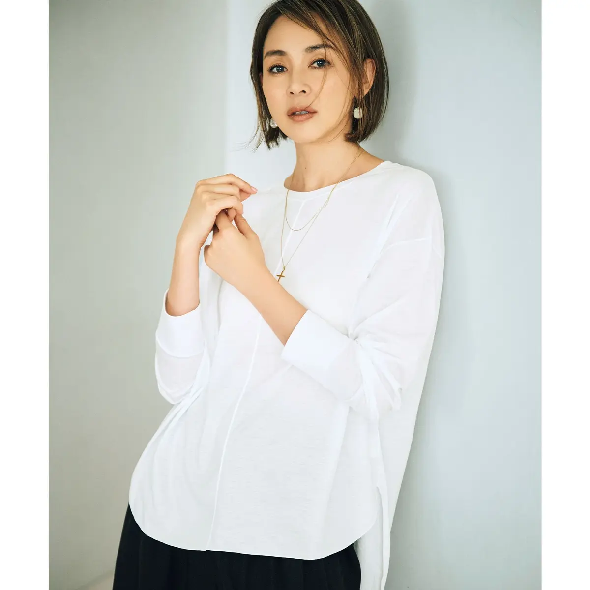 白い長袖カットソーを着た女性。肩口にドレープがあり、ゴールドのネックレスとピアスを着用。背景は淡いブルーの壁。