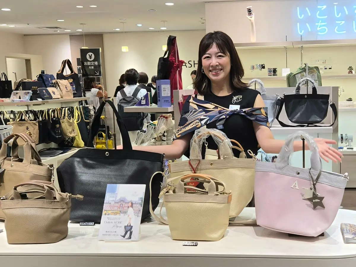デパートのバッグ売り場に立つ女性。黒のTシャツを着て、様々なデザインと色のバッグを身につけ、笑顔でこちらを見ている。背景には他の商品や買い物客が見える。