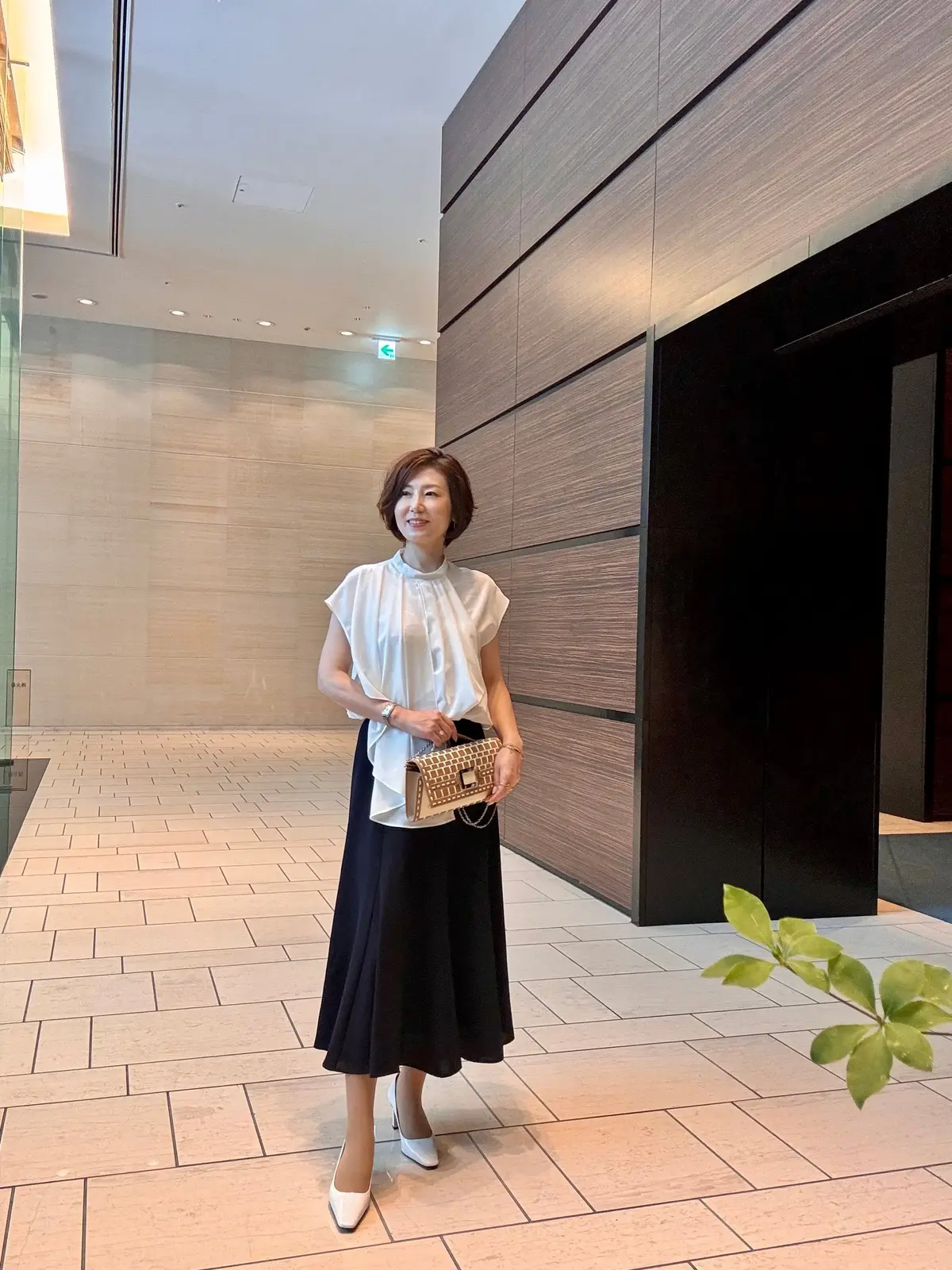 白い襟付きブラウスに黒のミモレ丈スカート、白いパンプスを着用した女性が立っている全身写真。背景はベージュと茶色のタイル張りの廊下。右手に小ぶりのバッグを持つ。