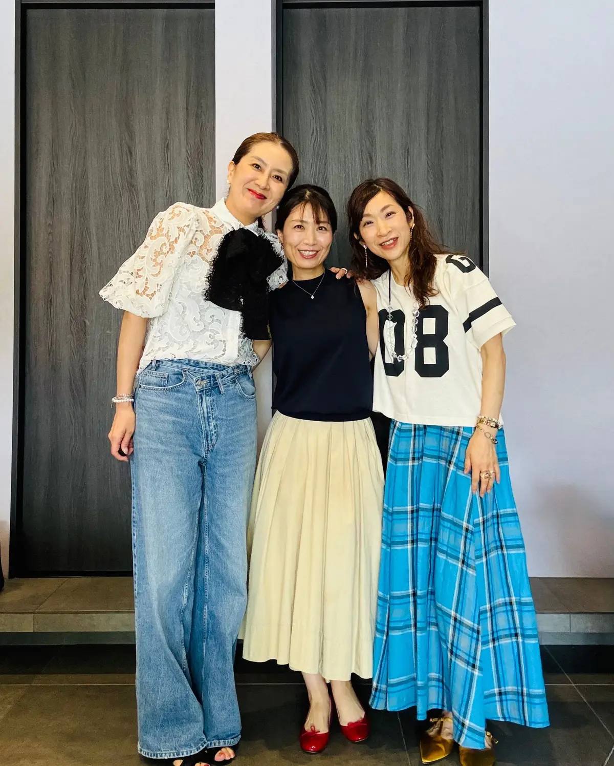 白のレース風トップス、黒のスカート、ジーンズを着た3人の女性が、木製のドアの前で並んで笑っています。中央の女性は黒のタンクトップとベージュのスカートを着て、赤いヒールを履いています。