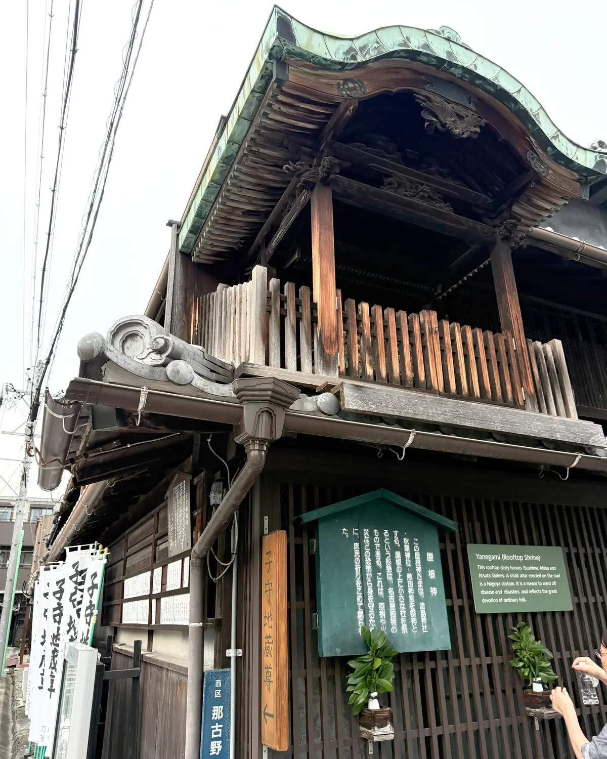 屋根の上に小さな神社が建てられた建物。木造で、緑色の屋根瓦と、装飾的な軒下がりの構造が特徴。建物前には説明書きと植木が置かれている。