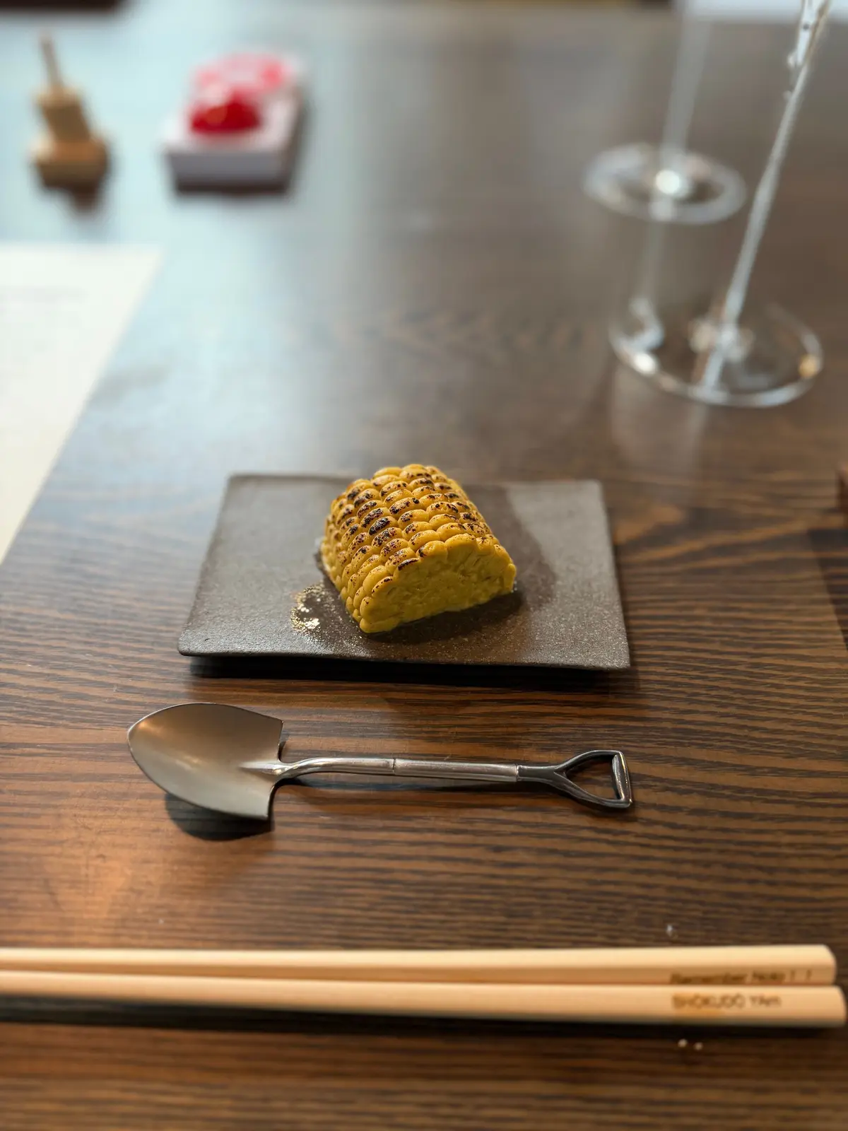 木製のテーブルに置かれた、焼き色のついたとうもろこし。黒い正方形の皿に盛り付けられ、シャベルのような形の銀食器と箸が添えられている。背景にはワイングラスと料理が見える。