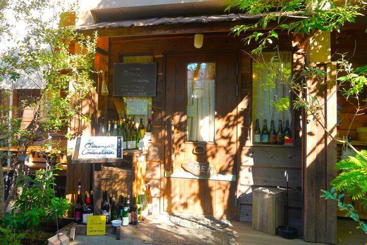 木造の小さな店舗の入り口。窓やドアにワインボトルが飾られ、手書きの看板が立てかけられている。緑の葉が茂り、木目調の建物と調和している。
