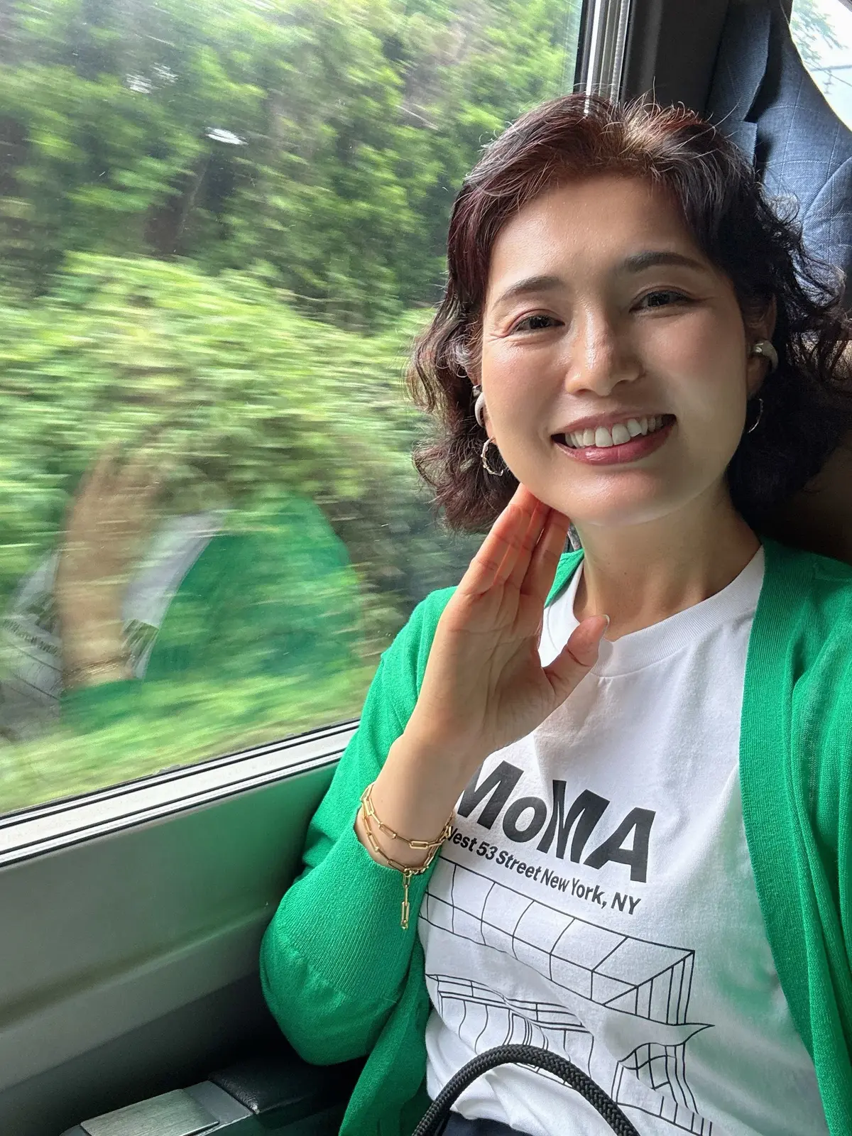 女性が緑の服を着て電車内で手を波し、緑の風景が見える窓のそばに座っている