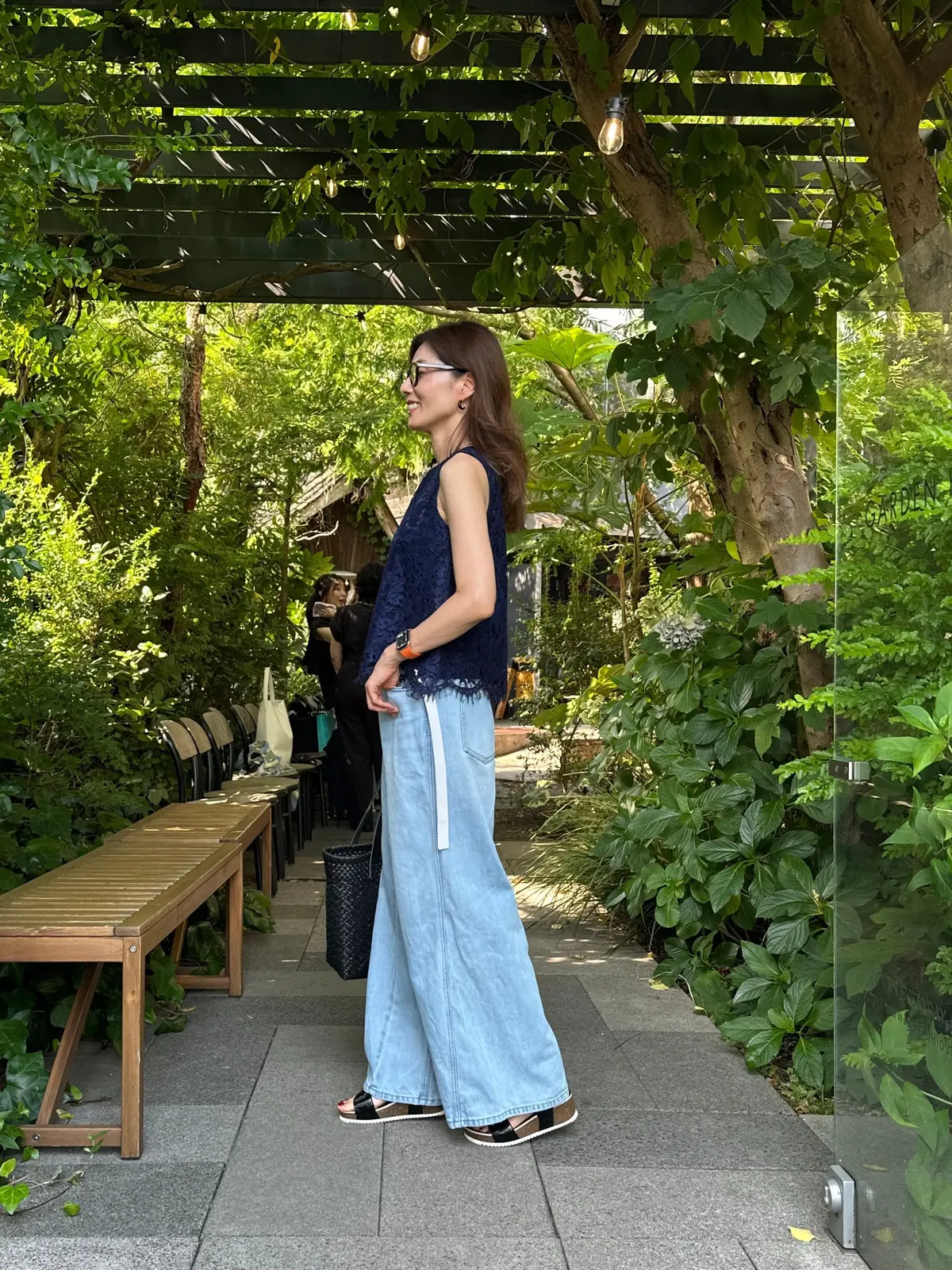 木陰のテラスで、女性がこちらを向いて立っている。濃い青色のトップスに、白いラインが入ったゆったりとしたデニムパンツを着用。黒縁の眼鏡をかけ、黒いサンダルを履いている。背景には緑豊かな植物と木々が見える。