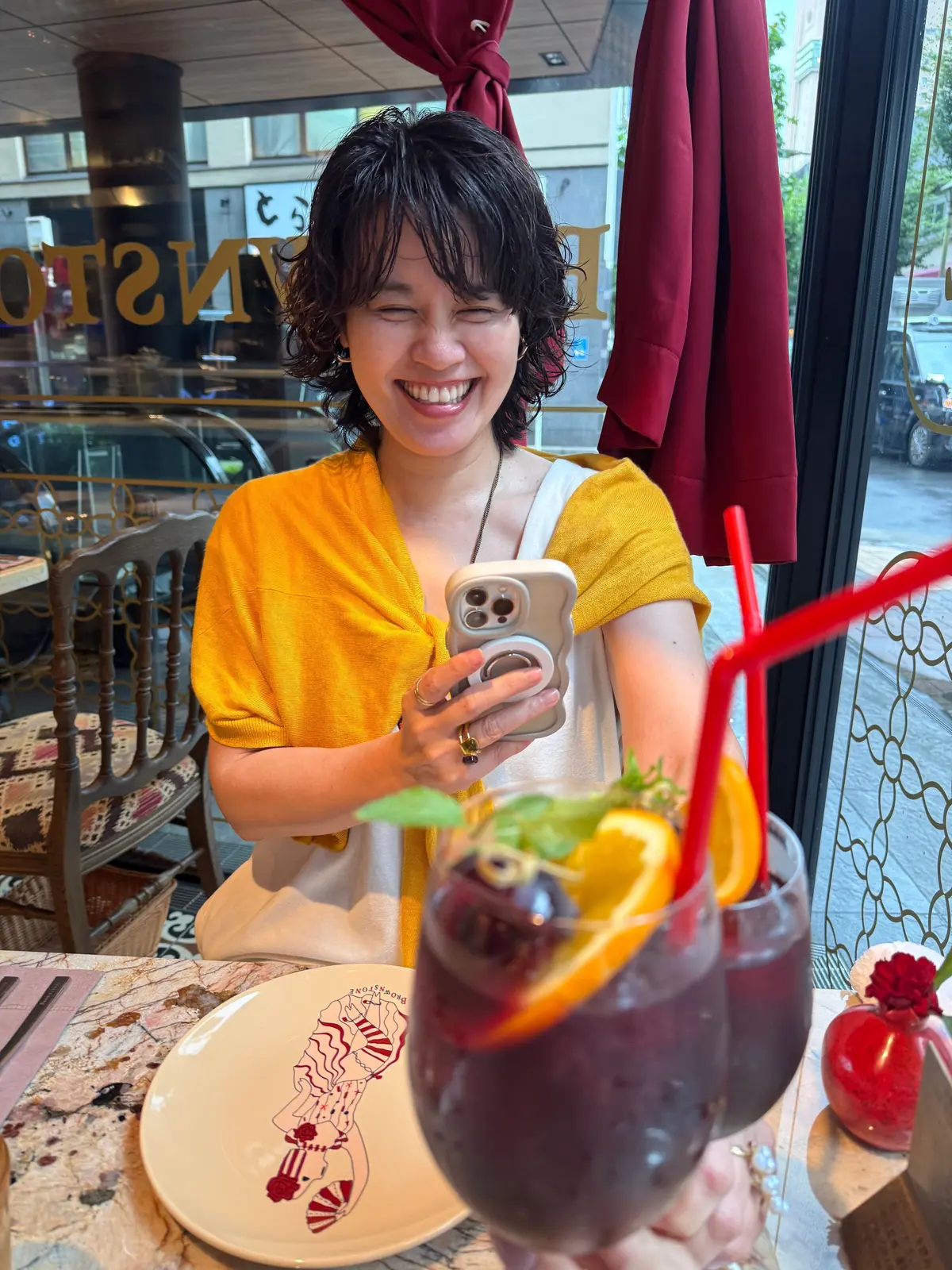 笑顔の女性が、オレンジ色のトップスを着てスマートフォンを見ている。テーブルには、赤い飲み物と、ロブスターのイラストが描かれたお皿が置かれている。背景には窓から街並みが少し見える。