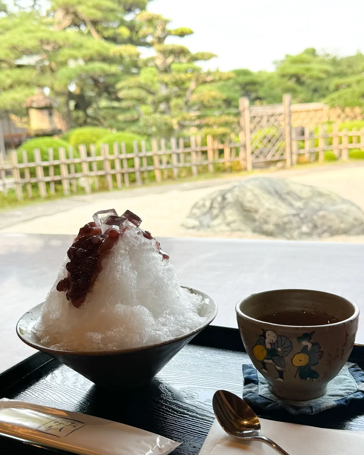 黒蜜と苺大福風のあんこが乗ったかき氷。背景には日本庭園が見える。茶碗に入ったお茶とスプーンも写っている。
