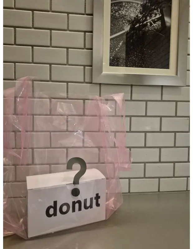 グレーのタイル壁の前に置かれた白い箱。箱には黒い文字で「? donut」と書かれ、ピンク色の透明な袋が被せられている。背景には、ドーナツの絵が飾られた額縁がある。