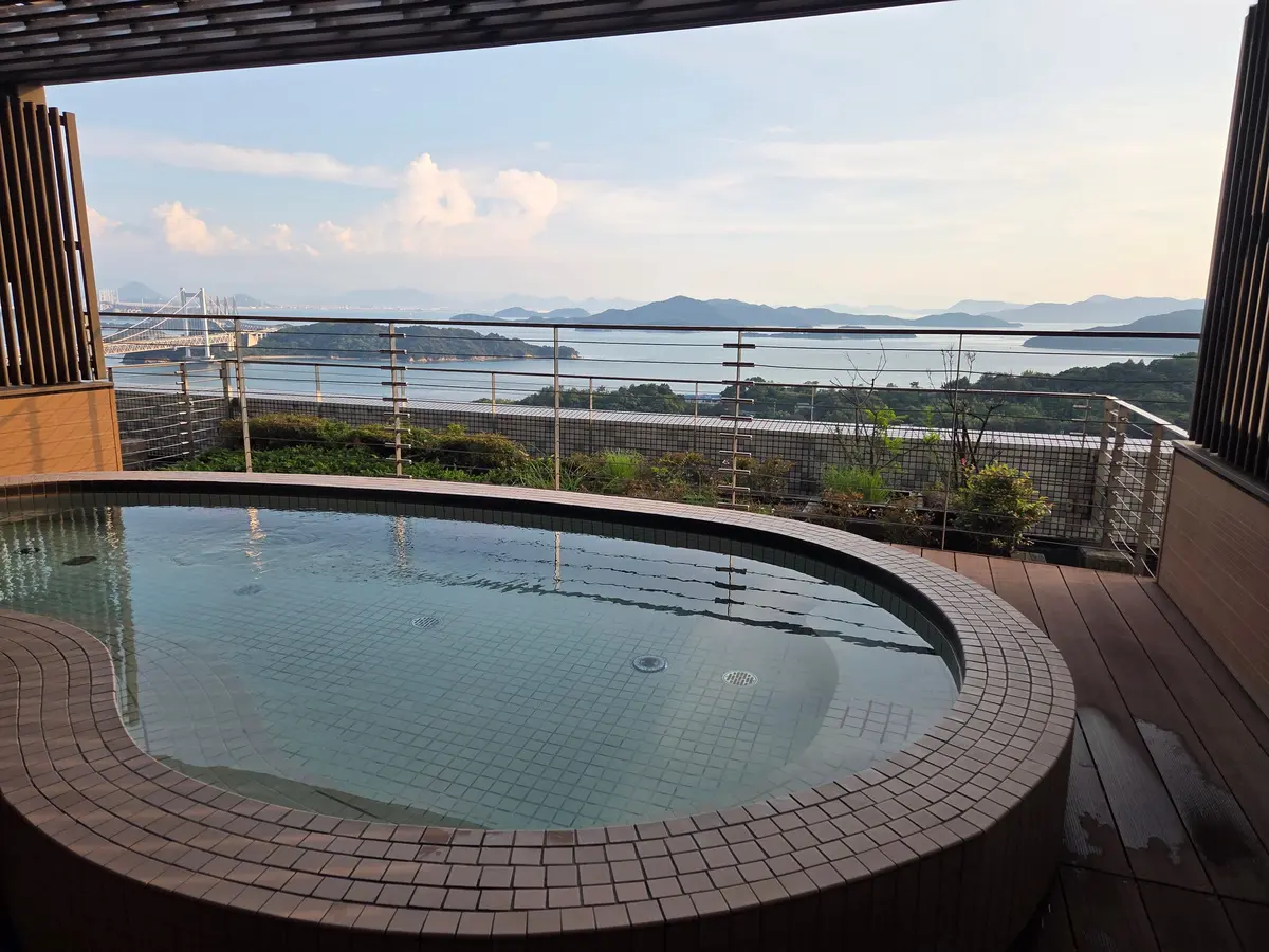 屋上露天風呂。丸い形状で、灰色のタイルで縁取られている。遠景には海と島々、橋が見える。青空に白い雲が浮かぶ。