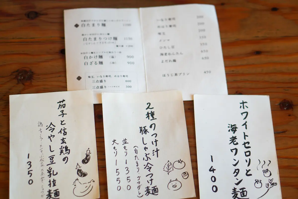 木製のテーブルに置かれた、複数のメニュー用紙。白文字で価格と料理名が記載された黒いメニューと、手書き風の文字で料理名と価格が書かれた白いメニューが混在している。右端には、白い背景に赤い文字で料理名と価格が記載されたメニューがある。