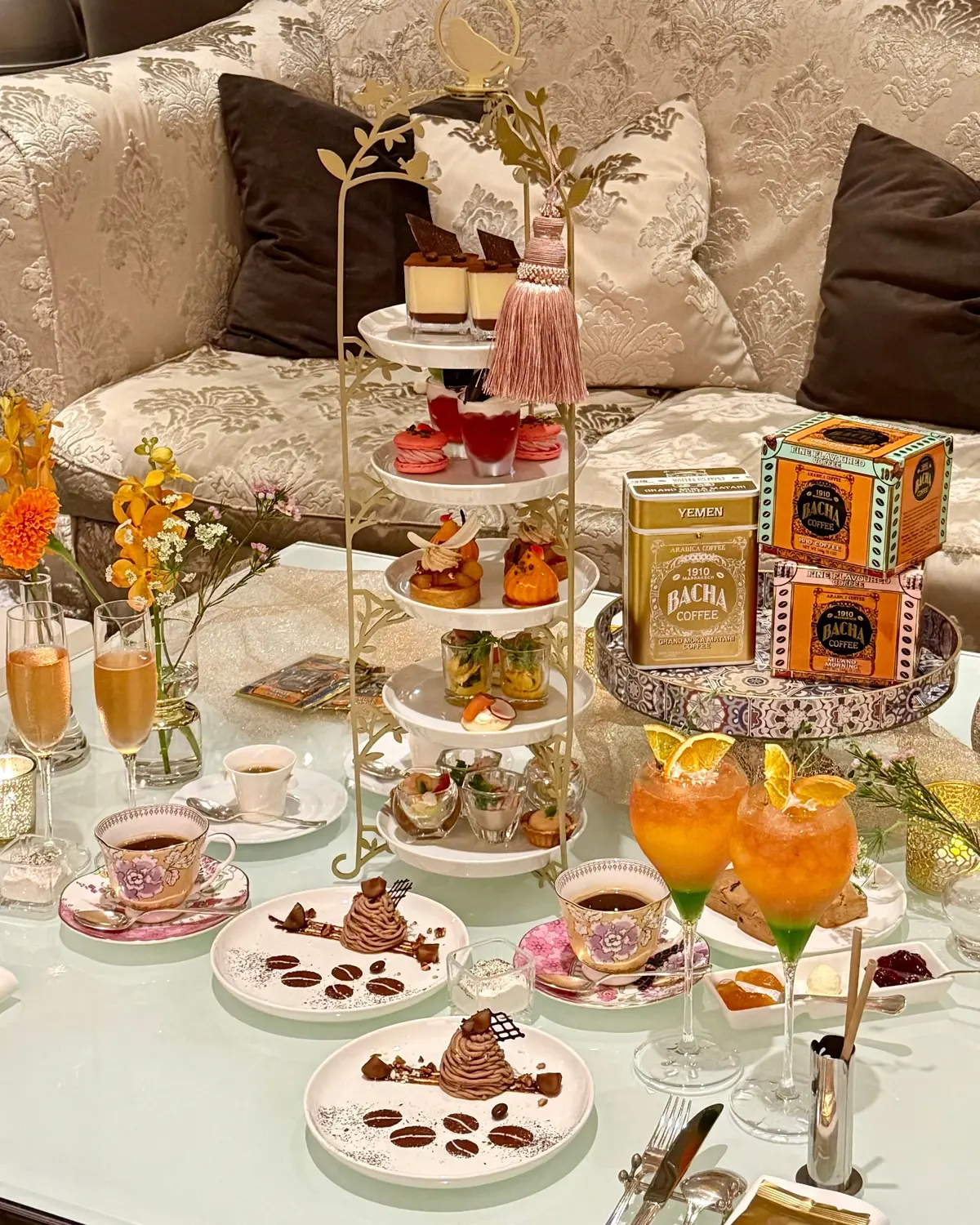 バッハコーヒーの箱と、3段のケーキスタンドにマカロンや焼き菓子が並ぶティーセット。グラスに入ったカラフルなカクテルや、コーヒー、小さなデザートも配置されている。背景には、装飾が施されたソファとクッションが見える。