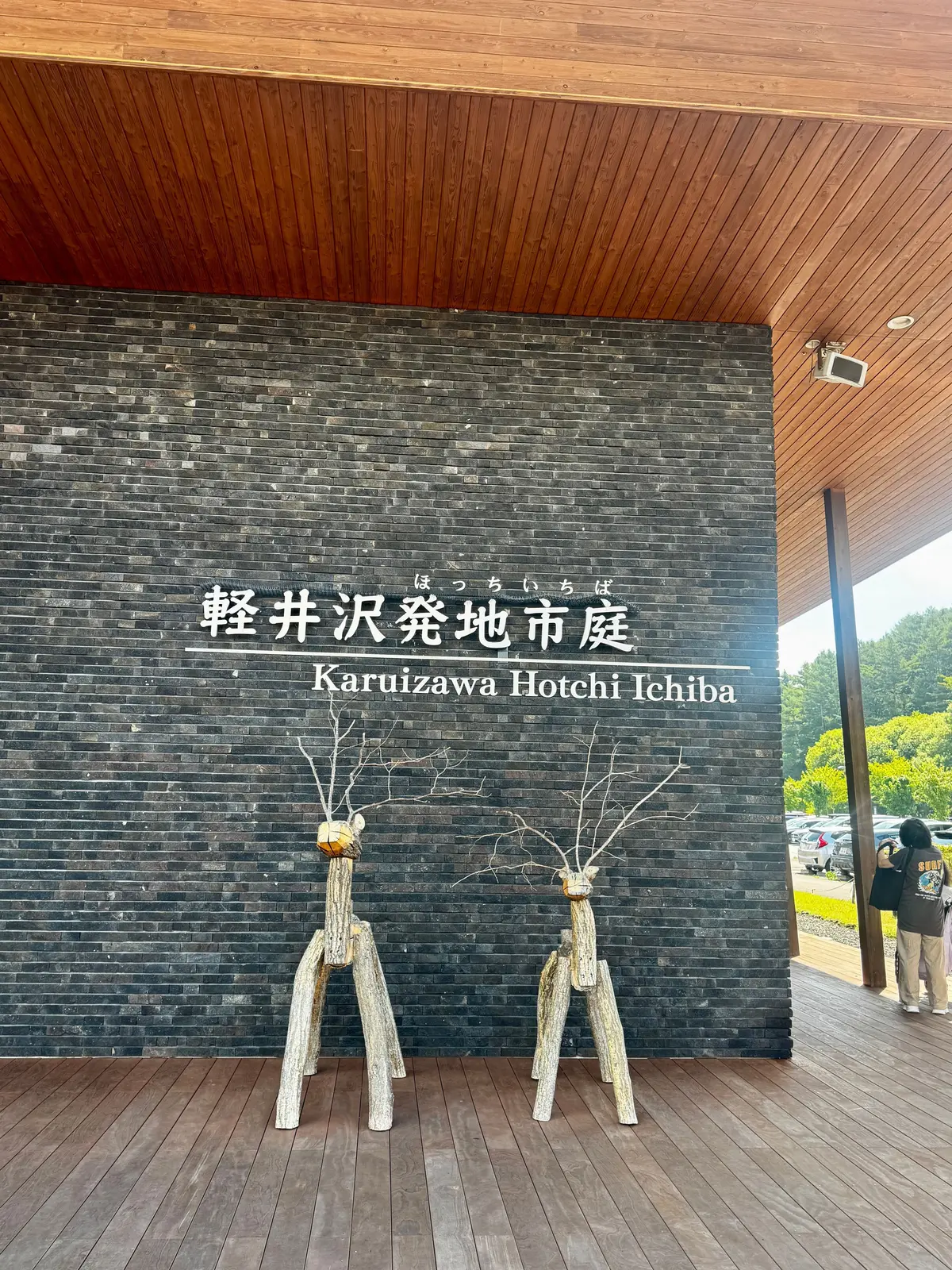 木造の庇の下、木製の柱が3本並び、その前に「KarUIzawa Hotchi Ichiba」の看板が設置されている。背景には緑の山々が広がる。