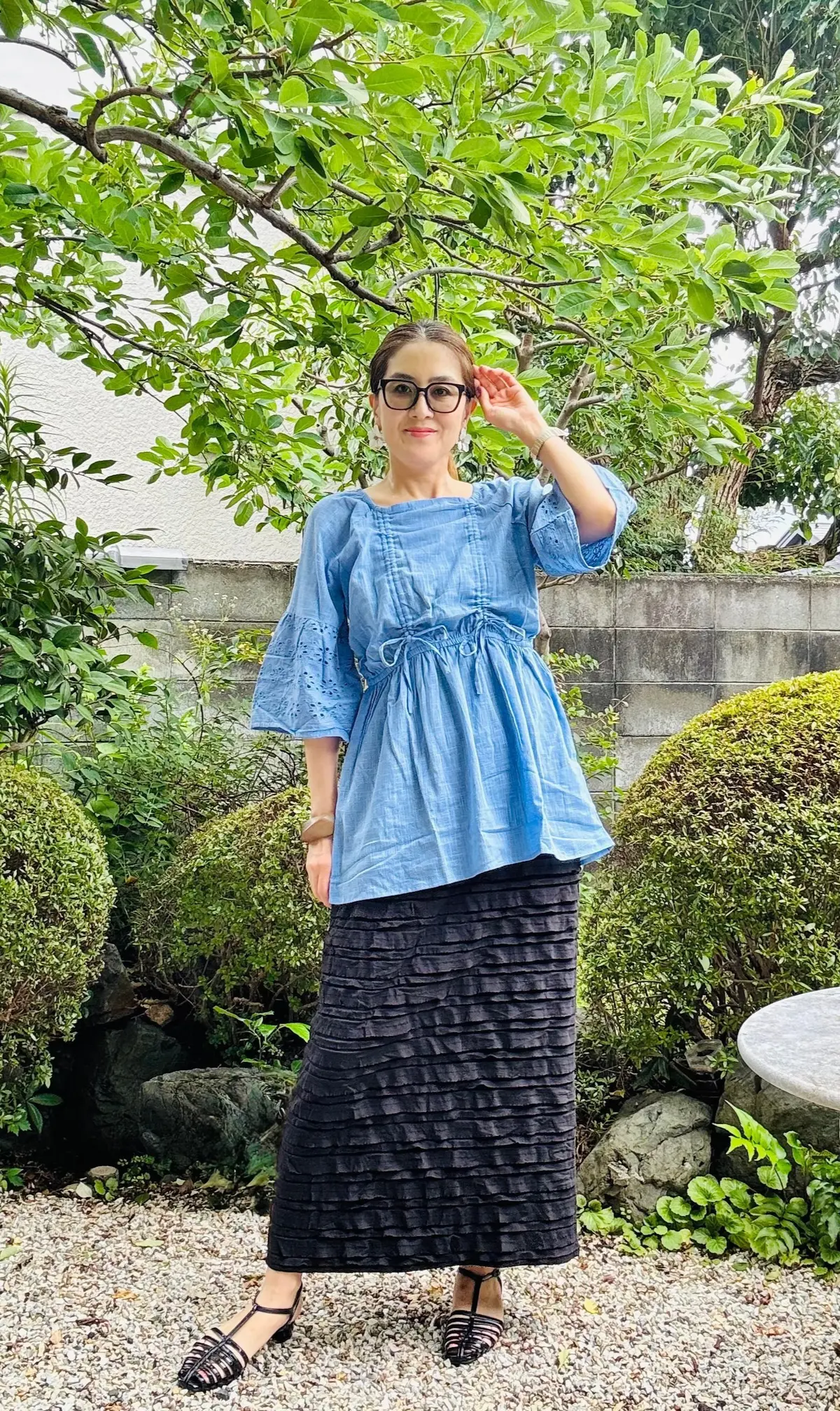 青いフレアスリーブのブラウスと黒いプリーツスカートを着た女性が、屋外の庭で手をかざしています。背景には緑の低木や木が並び、白い石のテーブルが置かれています。
