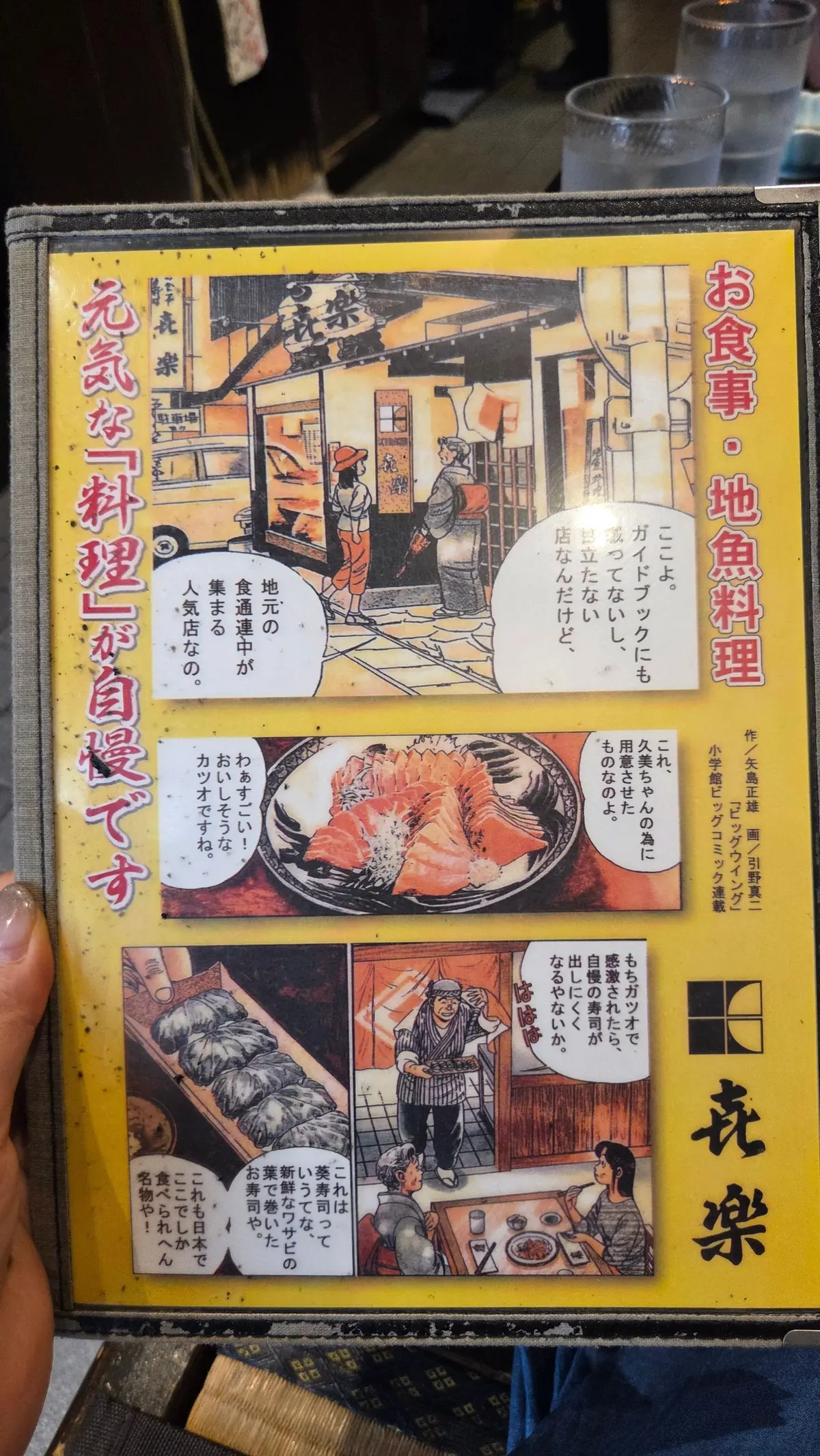 手塚治虫の漫画「火の鳥」のページ。鮮やかなオレンジ色の背景に、手塚のイラストと日本語のテキストが配置されている。中央には、カニ料理の写真と、それを囲むように漫画のコマが描かれている。
