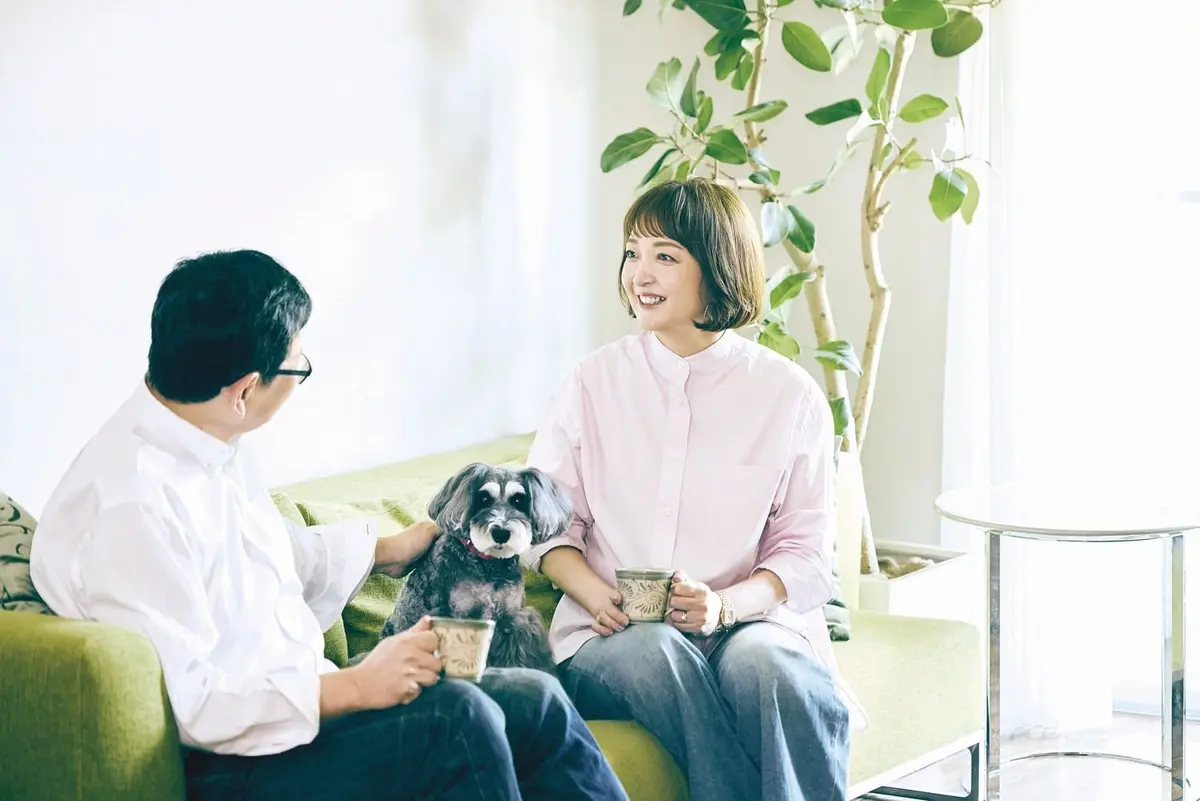 明るいリビングで、男女が犬と座って談笑している様子。男女はそれぞれ飲み物を手に持ち、犬は座って見つめている。背景には観葉植物が置かれている。