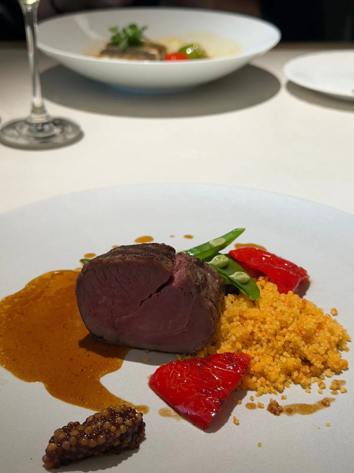 白磁皿に盛り付けられた、赤紫色の肉料理。肉の横には黄色いソース、赤と緑の野菜、茶色い粒状の食材が配置されている。背景にはワイングラスと別の料理が見える。
