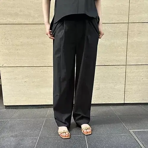 黒いワイドパンツと黒いトップスを着た人物が、ベージュの壁とグレーの大理石の床の前で、両手はポケットに突っ込んだ姿勢で立っている。足元にはオレンジ色のサンダルが履かれている。