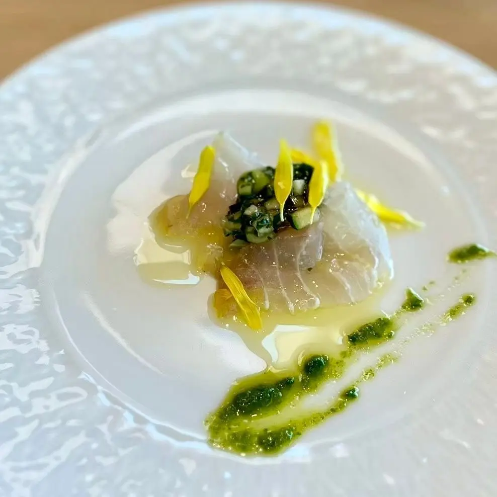 白いプレートに、薄い色の魚の切り身が中央に置かれ、緑の野菜のピクルスと黄色いエビのピースがトッピングされています。緑のソースがプレートに広がり、全体がエレガントな料理です。