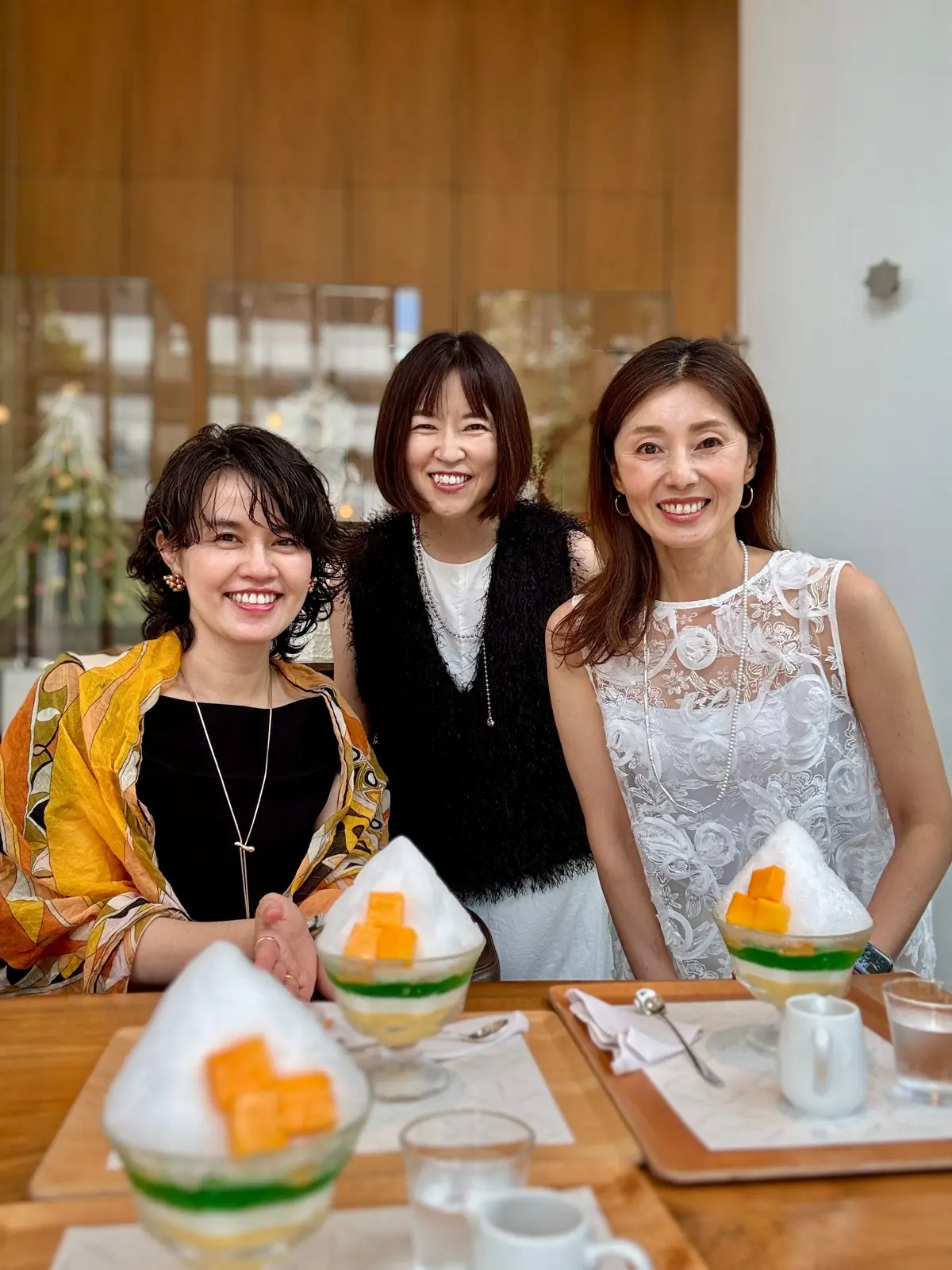 笑顔の3人の女性が、木製のテーブルを囲んで写っている。テーブルには、かき氷のようなデザートが置かれている。背景には、木製のドアと窓が見える。