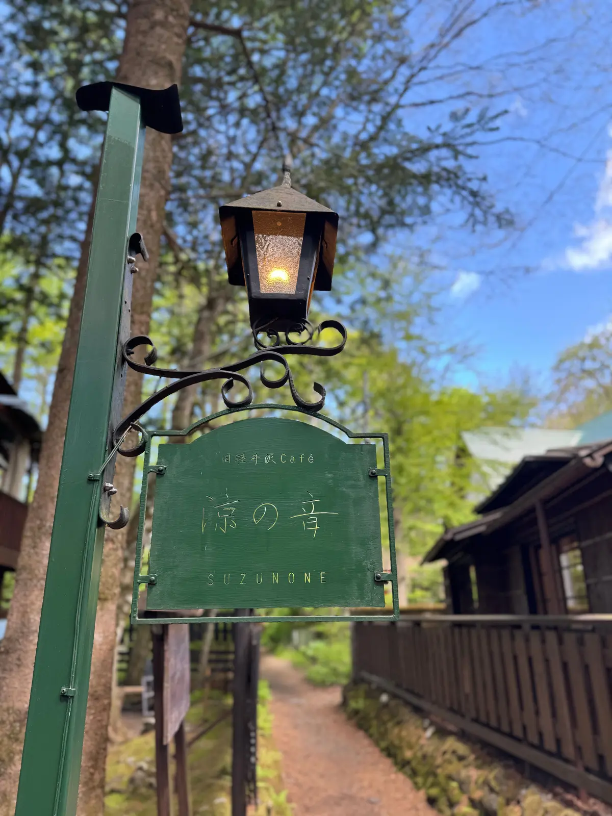緑の看板に「すずのねカフェ」の文字と「SUZUNONE」の表記があり、装飾された黒いランプが取り付けられた木製の柱に設置されている