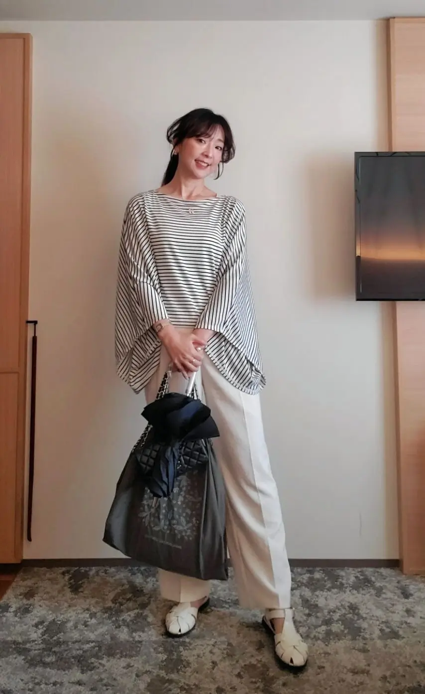 白いカーペットの上で、女性が笑顔で立っている。黒と白のボーダーのトップスに、白いゆったりとしたパンツを着用。グレーの大きなトートバッグを持ち、白い編み込みサンダルを履いている。背景には木製の家具とテレビがある。