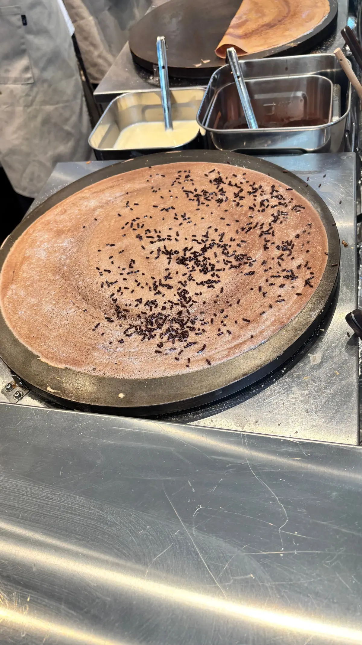 鉄板の上で焼かれるチョコレート色のクレープ。表面には細かい黒い粒状のものが散らばり、右上に生クリームが置かれている。