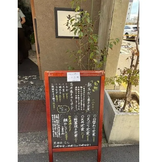 店舗の入り口に置かれた黒板のメニュー。手書きで料理名と値段が記載され、緑の植物が隣に植えられている。