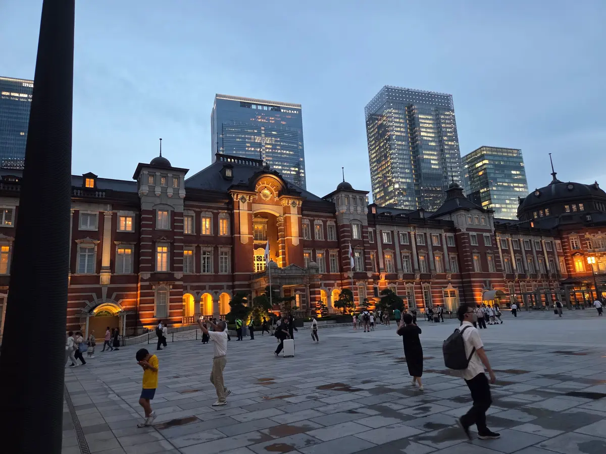 東京駅の赤レンガ造りの駅舎と、その背後にある高層ビル群。広場には多くの人が散策している様子。夕暮れ時の空が背景に広がる。