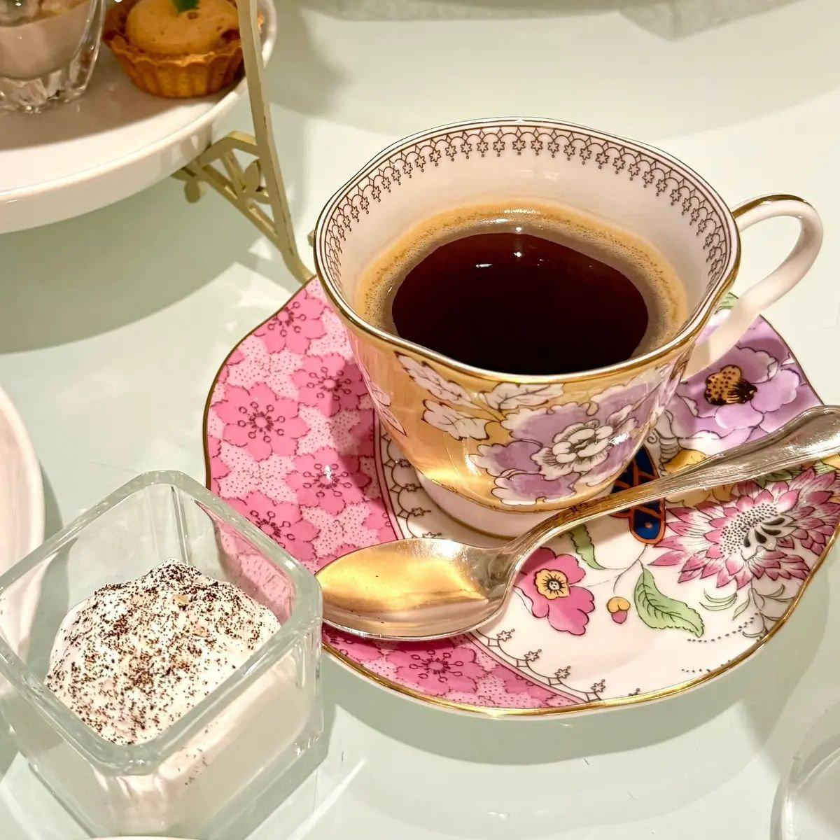 ピンクの花柄のソーサーに置かれたコーヒーカップと、その横の銀色のスプーン、砂糖入りの透明な容器が写っている。背景にはカップケーキが置かれた金色の台がある。