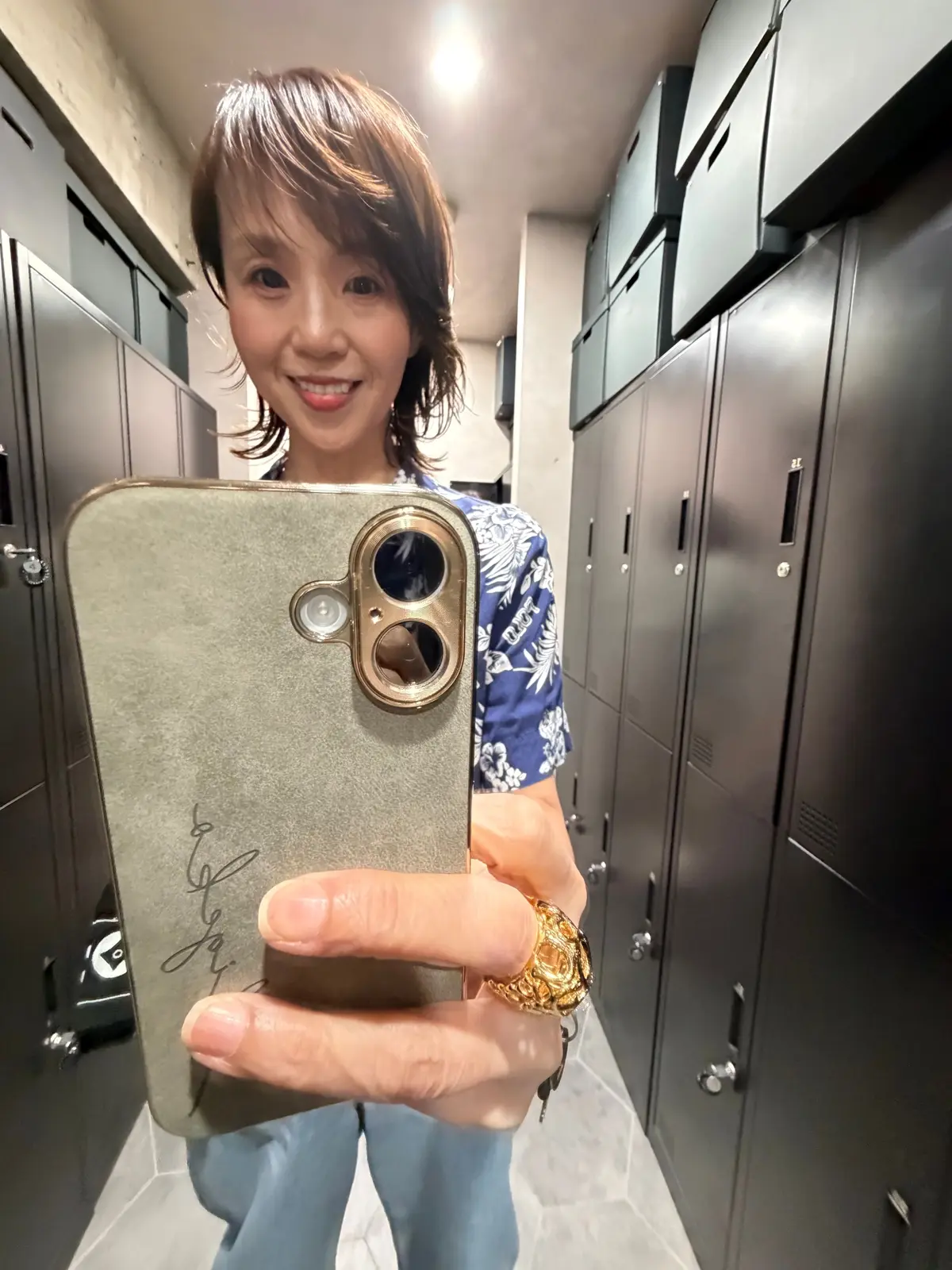笑顔の女性がスマートフォンで自撮りをしている全身写真。背景にはロッカーが並んでいる。女性は花柄のシャツとジーンズを着用し、ゴールドのネックレスと指輪をしている。