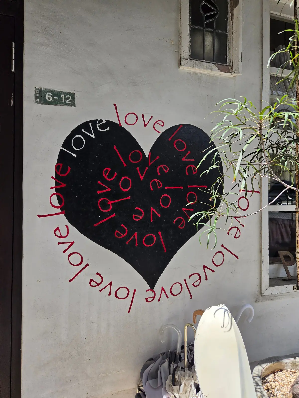 オフホワイトの壁に黒いハート型のステンシルアート。ハートの中には赤字で「love」の文字が散りばめられている。ハートの周囲にも赤字で「love」の文字が弧を描くように配置されている。壁の右側には窓と植物が写り込んでいる。