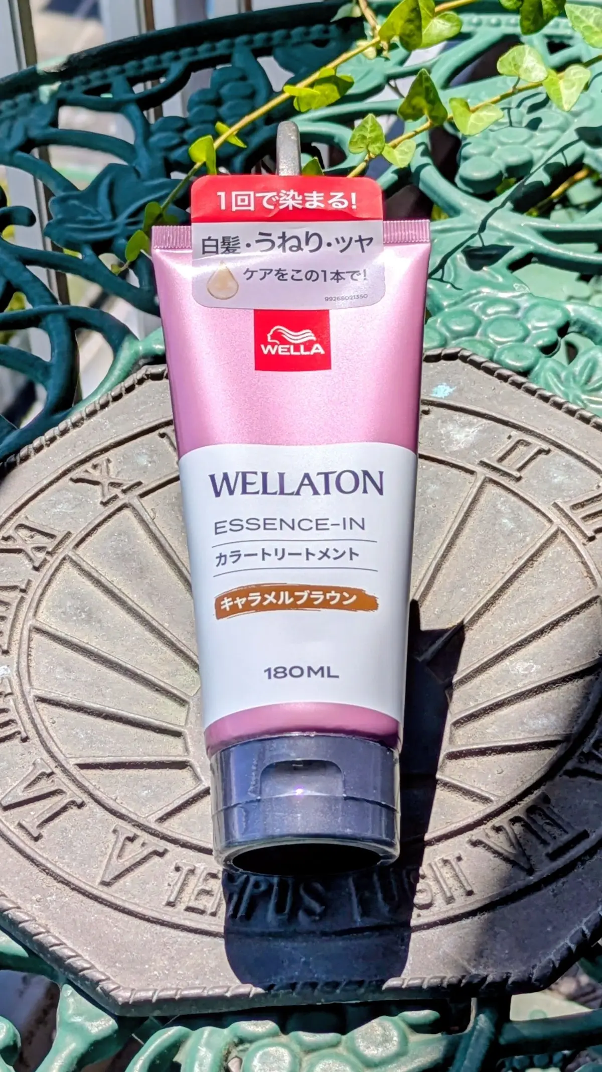 ピンクと白のチューブ型容器に入った、ウェラ（WELLA）のカラートリートメントメント。180mlで、キャメルブラウンの文字がデザインされている。背景には緑の葉が茂り、古びた金属製のベンチが置かれている。