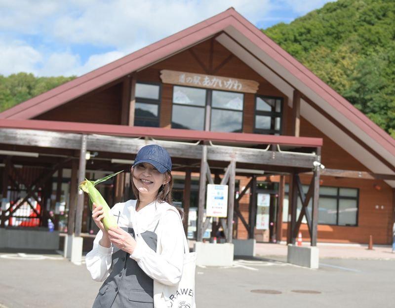 帽子を被った女性が、木造の道の駅の前に立って、とうもろこしを持っている。白いシャツにグレーのビスチェ、白いバッグを身につけている。背景には緑の丘が広がる。