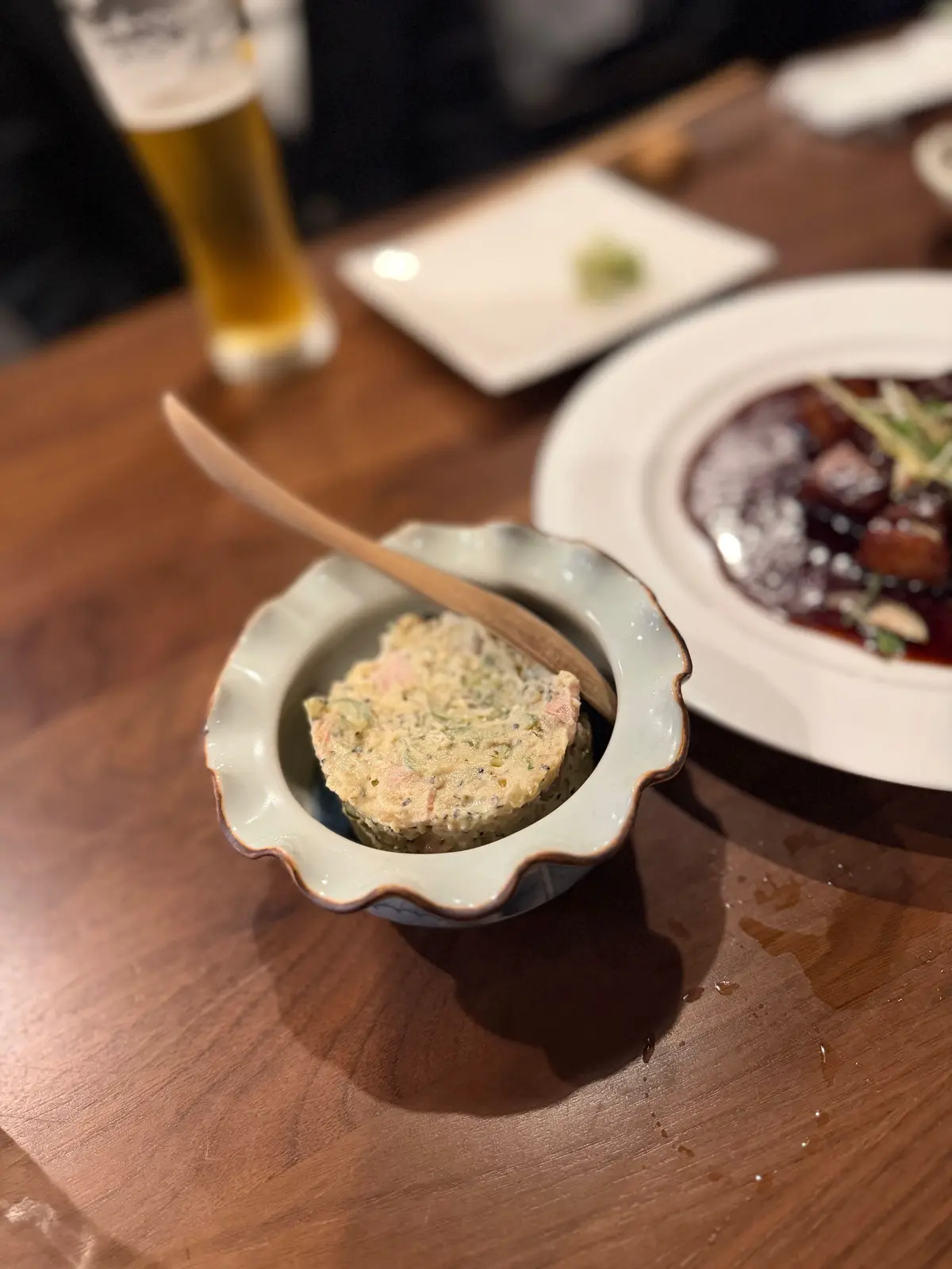 木製のテーブルに置かれた、青い器に入った淡い黄色の料理と、隣の白い皿の赤いソースが目立つ料理、そして背景のビールグラス。