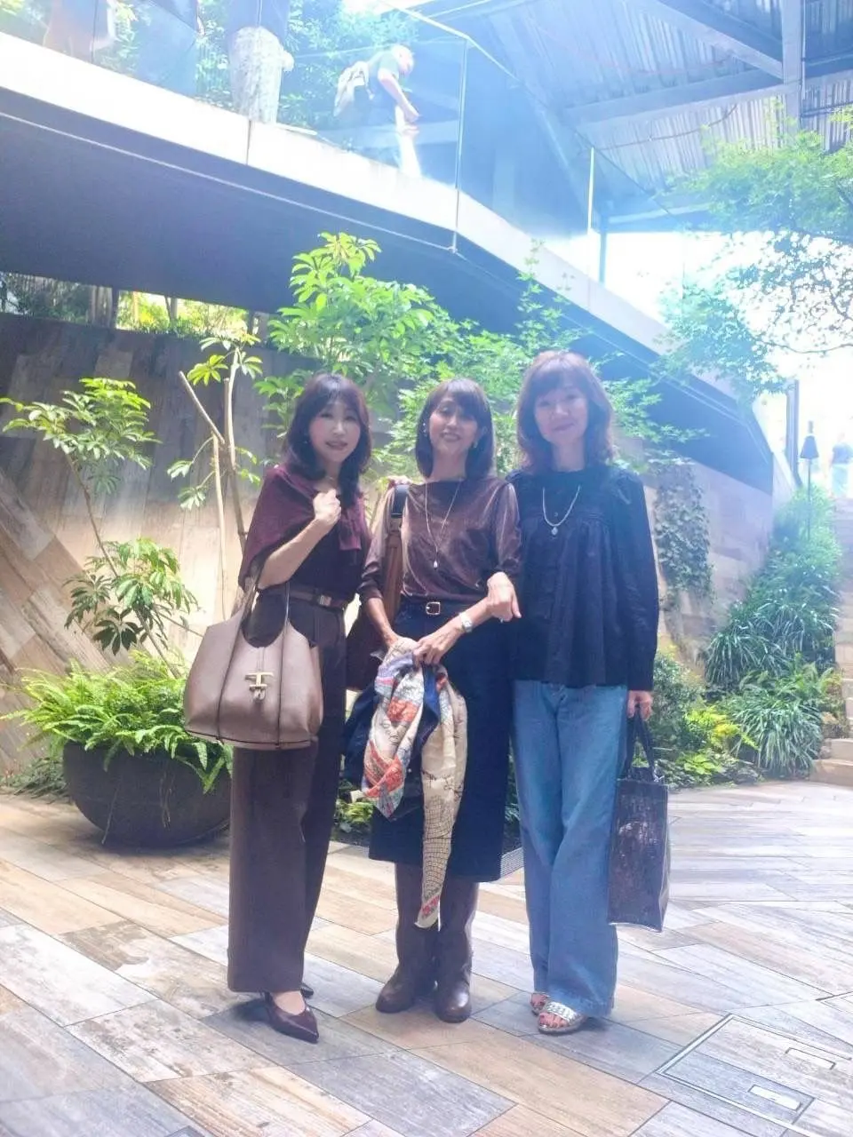 3人の女性が屋外の木製デッキで並んで立っている写真。左から、茶色のワンピースを着た女性、黒いトップスとスカート姿の女性、黒いトップスとジーンズ姿の女性。それぞれがバッグやアクセサリーを持ち、笑顔を見せている。背景には緑の植物と建物の構造物が見える。