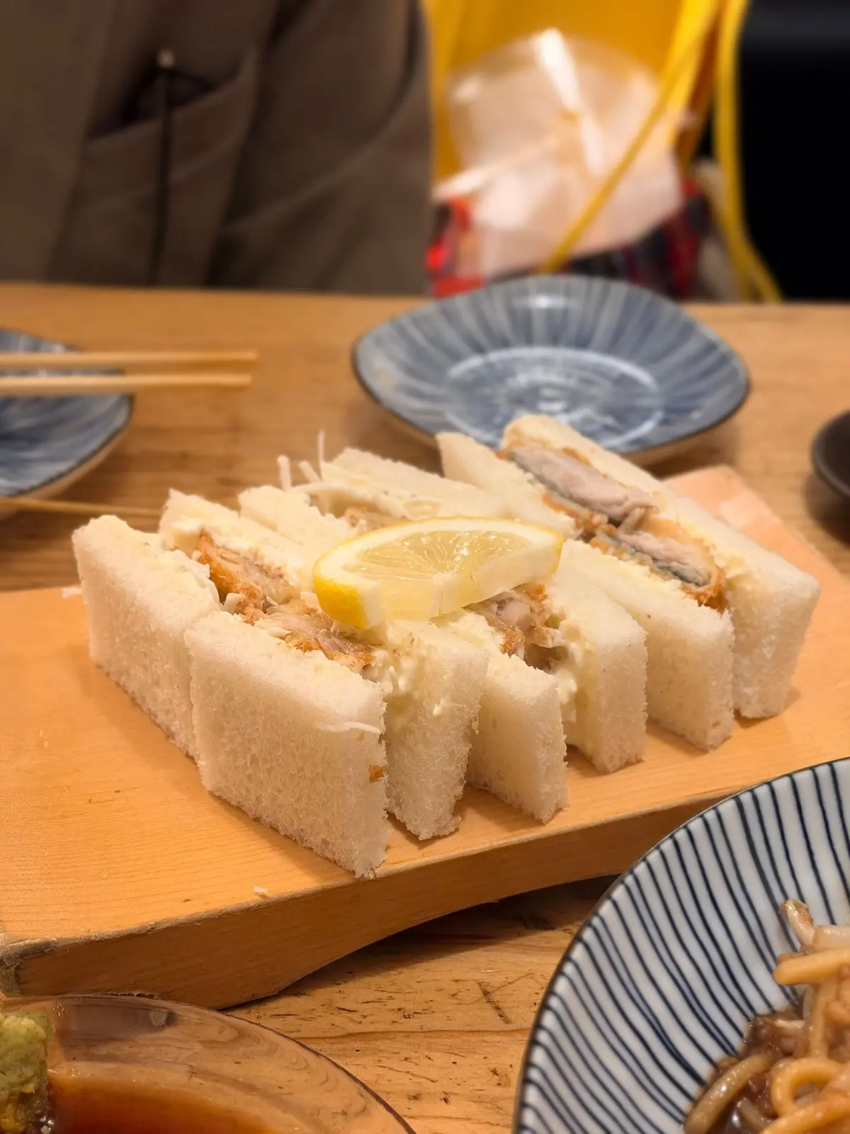 木製の板に並んだ、白くて四角いサンドイッチ。具材は魚肉と思われ、レモンが添えられている。背景には食器や人物がぼやけて見える。鯖サンド