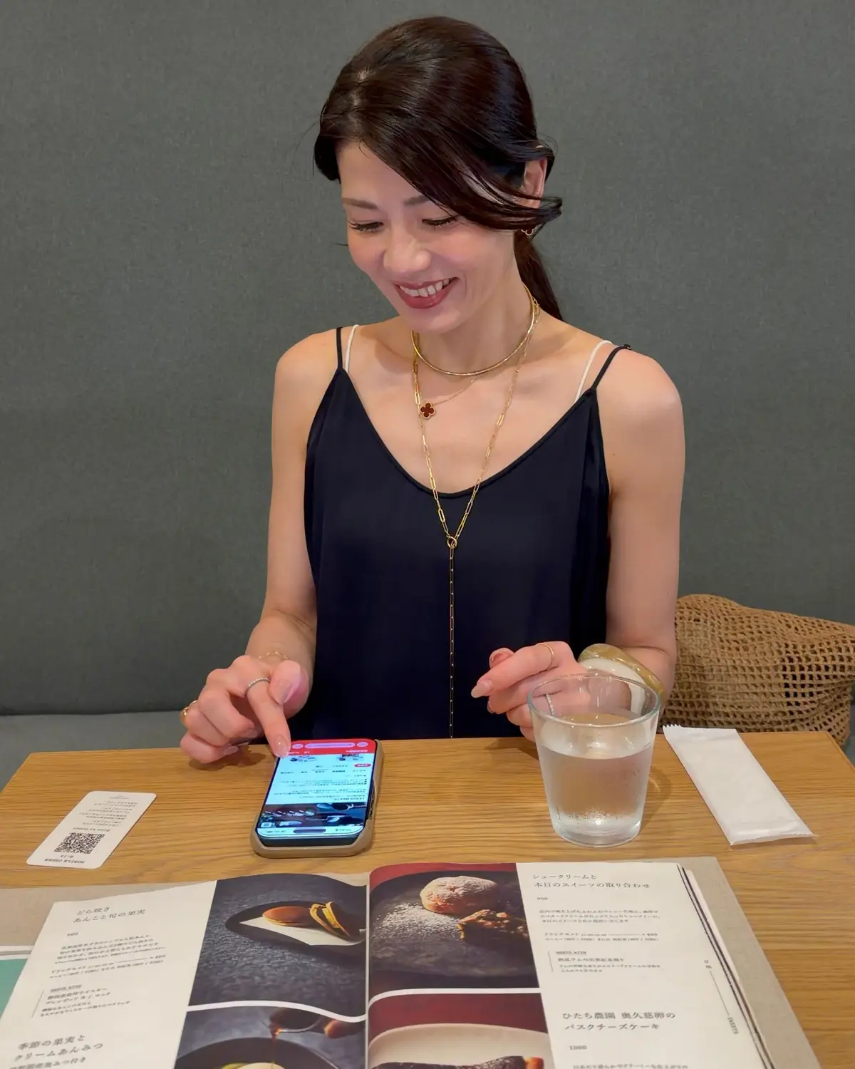 レストランでメニューを見ながらスマートフォンを操作する女性。黒いトップスにゴールドのネックレスを着用し、笑顔を見せている。テーブルにはグラスと日本語のメニューが置かれている。背景はグレーの壁。