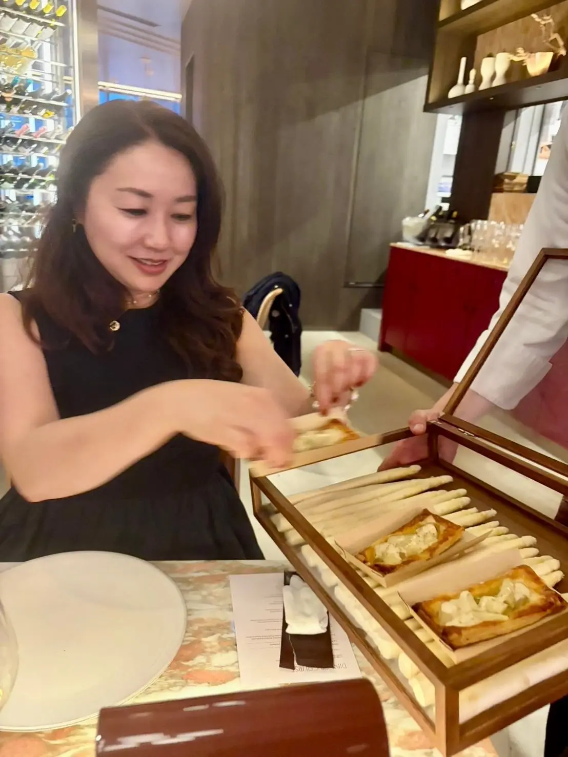 レストランのテーブルに、女性がパンのプレートを受け取る様子。プレートには、チーズをトッピングした焼き菓子と細長いパンが並び、背景にはワインのボトルが並ぶ棚が見える。