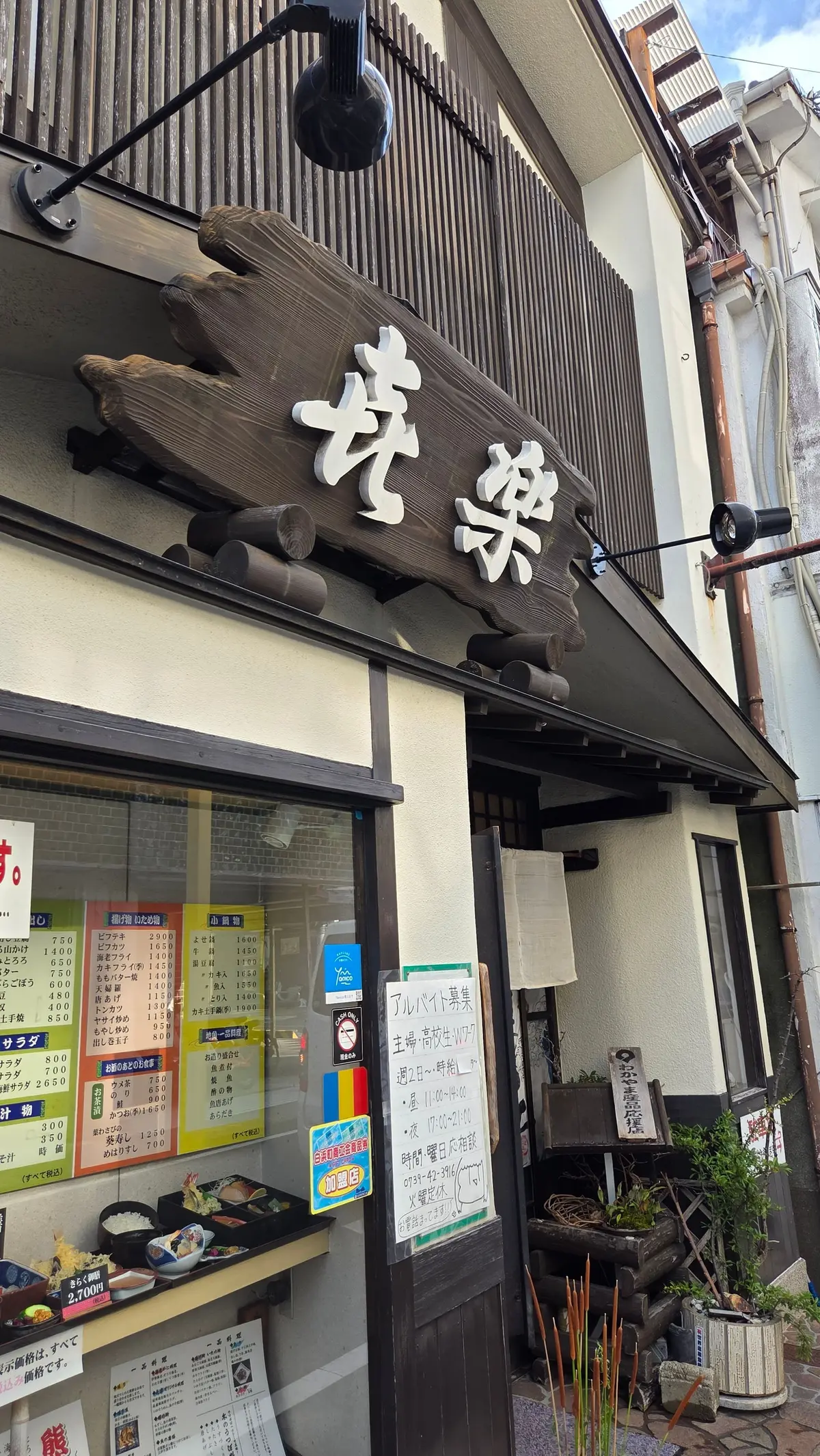 木造の看板が目立つ、日本の飲食店の外観。看板には白文字で「我楽亭」とあり、伝統的な装飾が施されている。窓にはメニューや案内板が貼られ、右には植栽が置かれている。