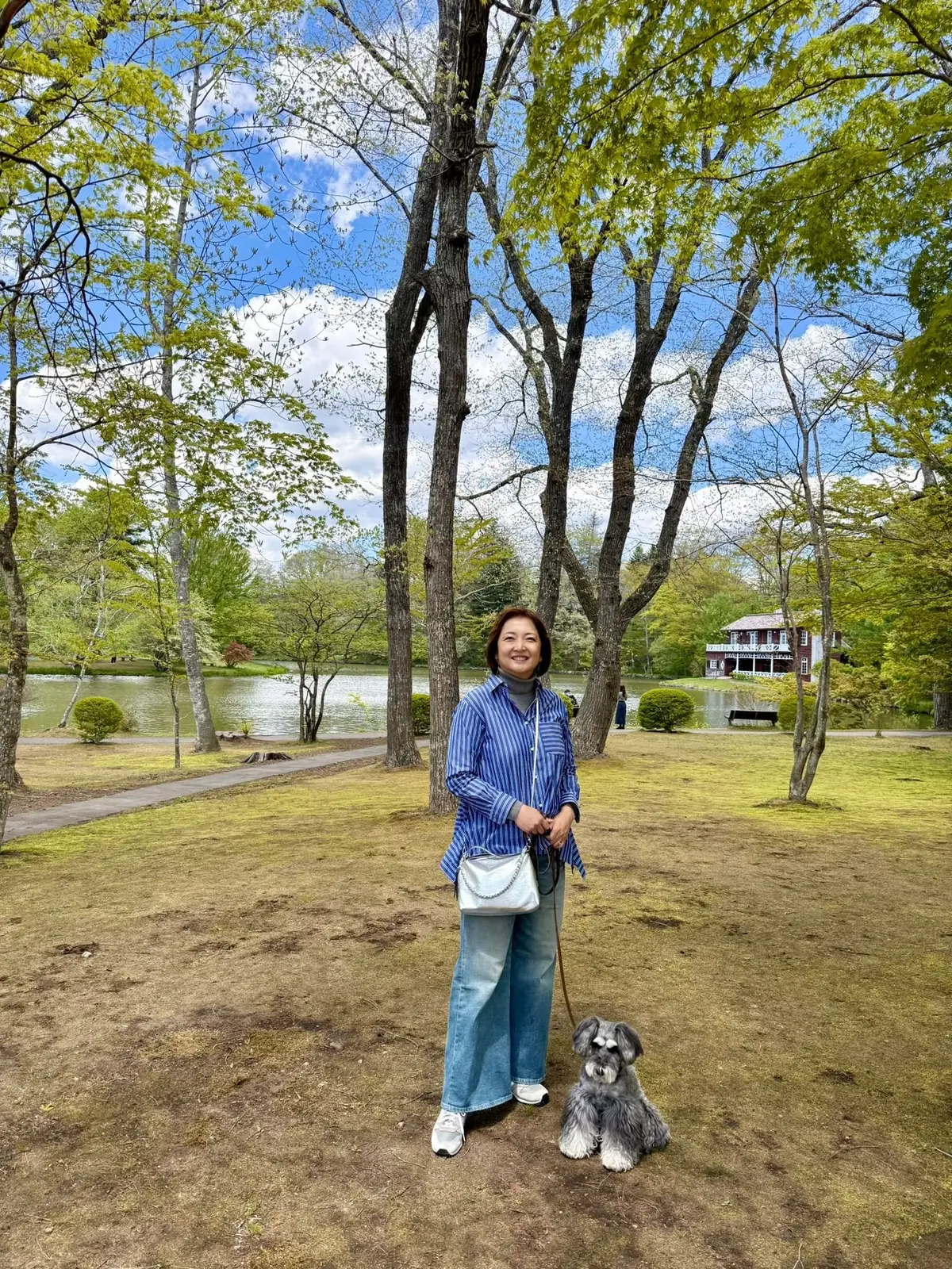青空の下、芝生の上に女性と小型犬が並んでいる。女性は青いストライプのシャツとジーンズを着て、白いショルダーバッグを持っている。背景には池と木々が広がり、赤い建物が見える。