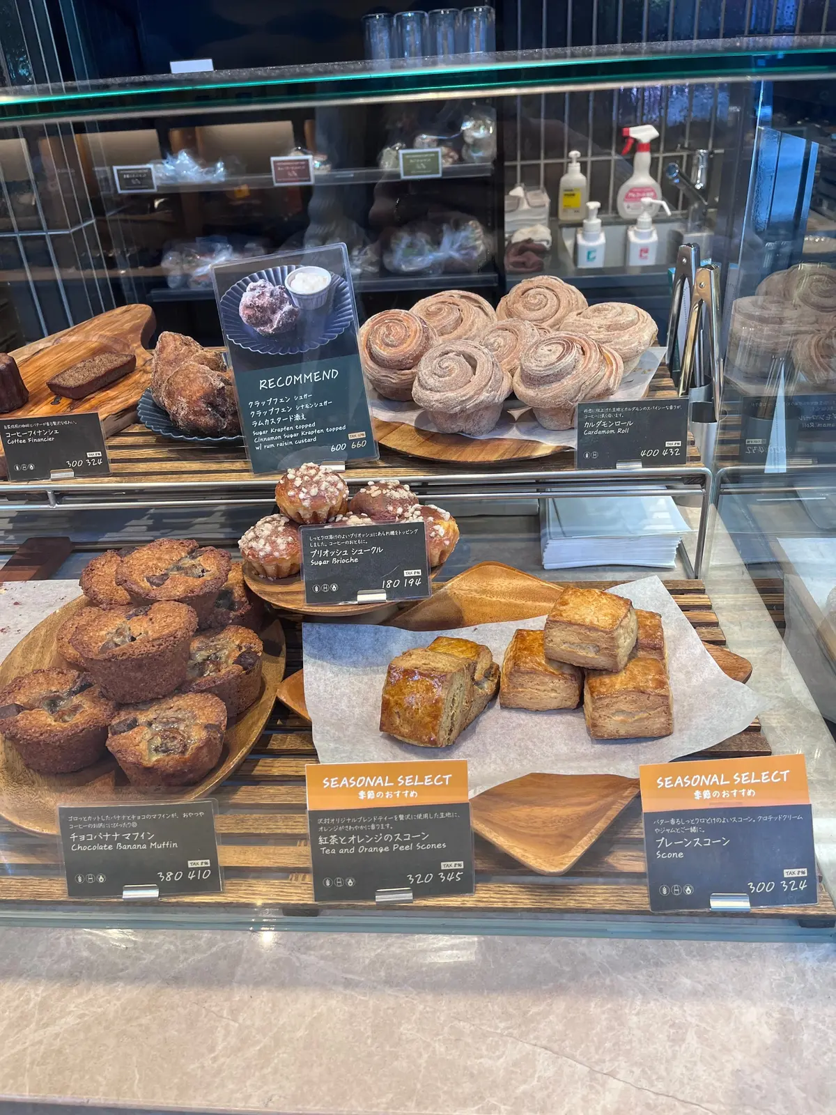パン屋のショーケースに並んだ様々な焼き菓子。中央には「RECOMMEND」と書かれた立て札があり、ロール状のパンや宝石のような焼き菓子、カップに入ったプリンなどが並んでいる。下段にはマフィンやスコーンが並び、季節限定のメニューも表示されている。
