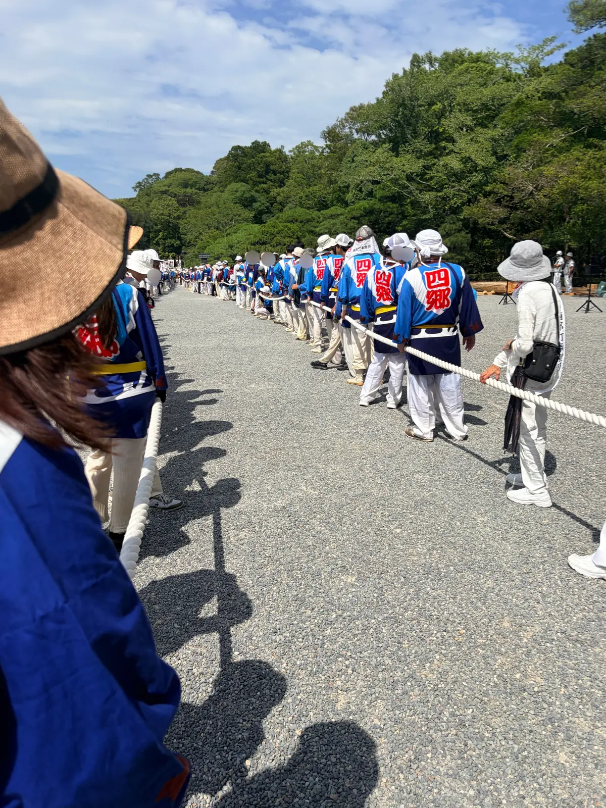 青い衣装をまとった人々が、白いロープで区切られた道を列をなして歩いている様子。背景には緑豊かな木々が見える。