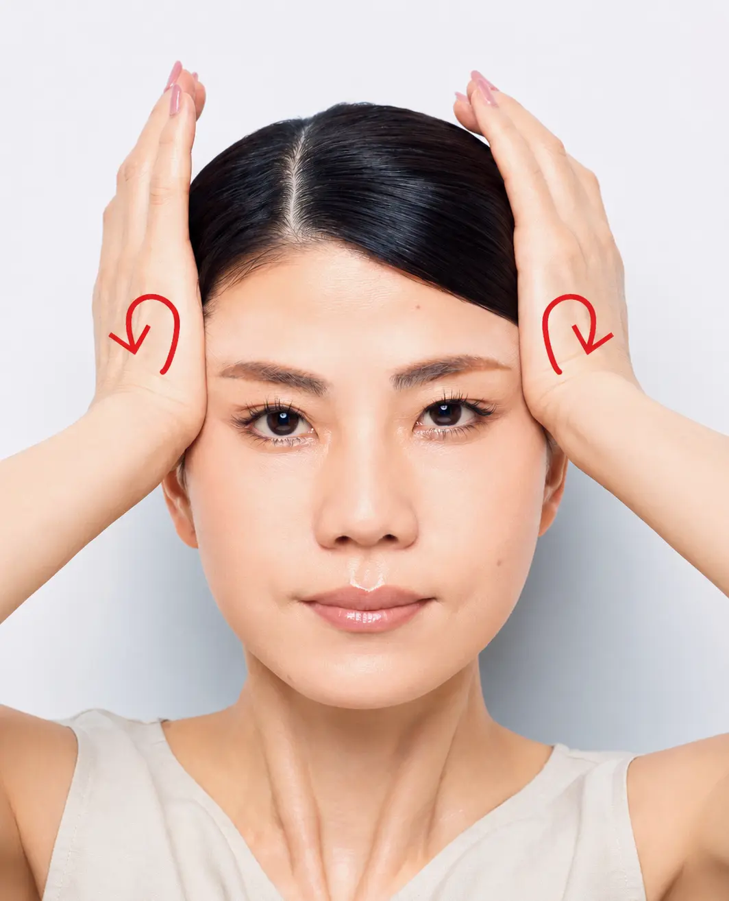 白い背景をバックに、タンクトップを着た若いアジア系の女性が正面を向いている。両手でこめかみを持ち、目元にはアーチ状の仕草をしている。ダークブラウンの髪を後ろでまとめ、自然なメイクをしている。