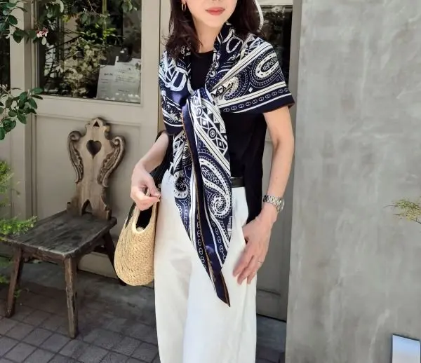 女性が青と白のパイソン柄スカーフを首に巻き、黒のトップスとベージュのワイドパンツを着用。左手には編みかごバッグを持ち、木製の椅子と窓のそばに立っている。