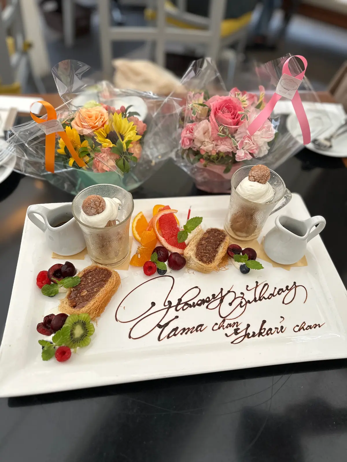 白い長方形の皿に、誕生日のお祝いメッセージ「Happy Birthday Mama chan, Rikari chan」がチョコレートソースで書かれている。