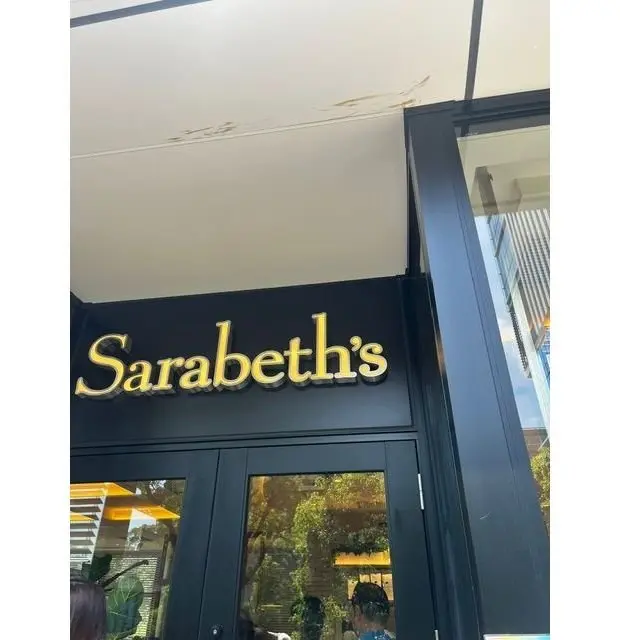 黒い看板に金色の文字で「Sarabeth's」と書かれたレストランの入り口。窓に緑の植栽が見える。