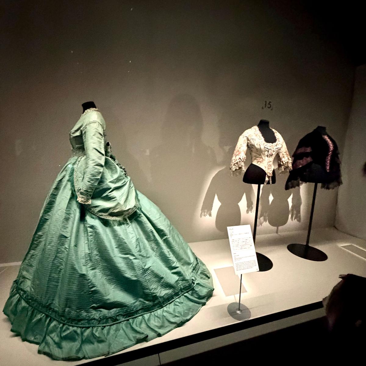 博物館の展示ケースに展示された、19世紀の女性用ドレス3点。左は淡い緑色の広がりスカートと長袖のドレス、中央は白地に金糸の刺繍が施されたドレス、右は黒いベルベットのドレス。それぞれマネキンに着せられ、説明書きが置かれている。