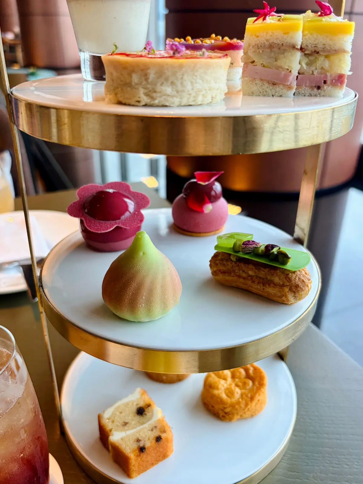 金色の支柱を持つ3段のケーキスタンドに、様々な軽食とスイーツが盛り付けられている。一番上段にはサンドイッチと小さなケーキ、真ん中段には花形のタルトや緑色の果物、シュークリーム、一番下段にはスコーンとパウンドケーキが並んでいる。
