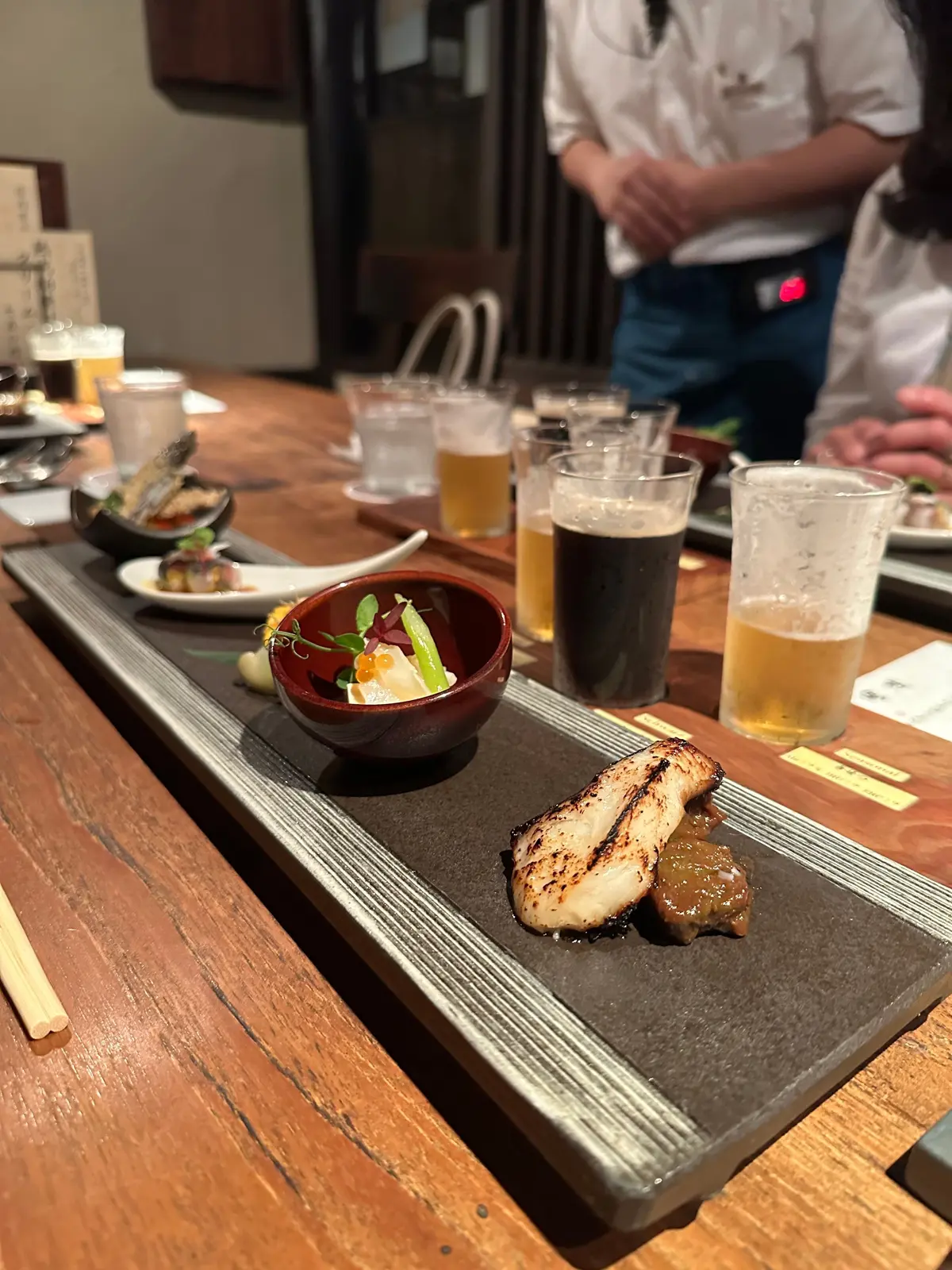 木のテーブルに置かれた料理とビール。焼き魚の盛り合わせ、小さな赤いボウルに入ったサラダ、複数のビールグラスが並ぶ。
